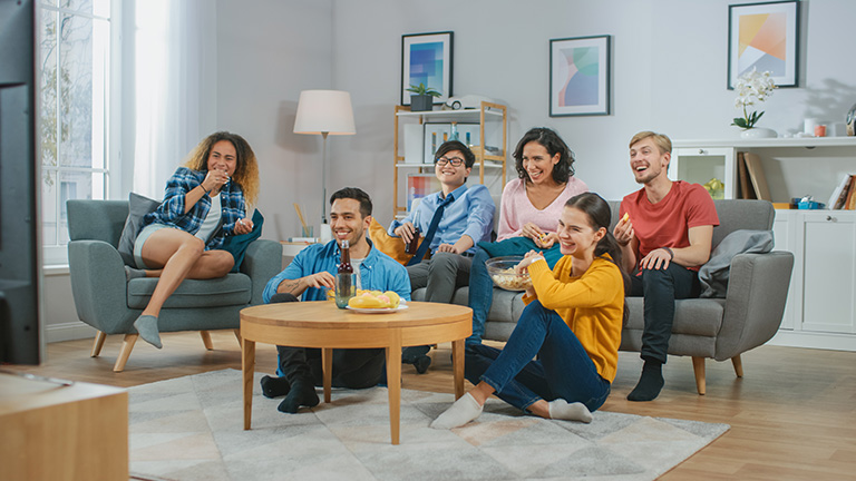 At Home Diverse Group Friends Watching TV Together, Eating Snacks and Drinking Beverage. Watching Comedy Sitcom or a Movie, Laughing and Having Fun Together.